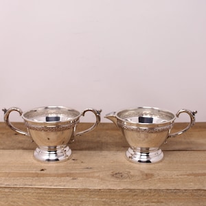 May include: A silver creamer and sugar bowl set with ornate detailing around the rim. The set is on a wooden surface.