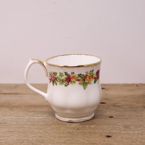 May include: White ceramic mug with a floral garland design featuring roses and greenery. The mug has a gold rim and handle accents. The mug sits on a wooden surface.