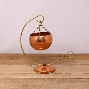 May include: A copper hanging planter with a brass stand. The planter is round and has a shiny finish. The stand is curved and has a round base.