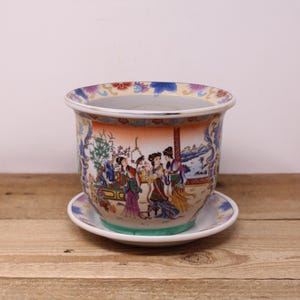 Peut inclure: Un pot de fleurs décoratif en céramique avec sa soucoupe assortie. Le pot présente une illustration colorée et détaillée de personnages en vêtements traditionnels, sur fond de paysage. Le bord et la soucoupe ont des motifs floraux. Le pot mesure environ 15 cm de diamètre.