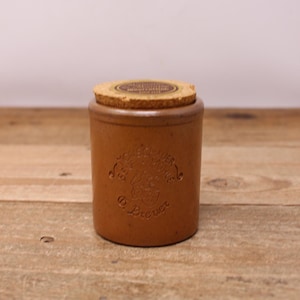 May include: A cylindrical, brown ceramic jar with a cork lid. The jar has embossed text and a design on the front. The cork lid is circular and fits snugly on top. The jar is sitting on a wooden surface.