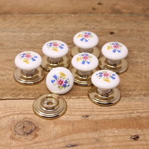 May include: Eight white ceramic door knobs with gold bases and floral designs in shades of pink, blue, and yellow.