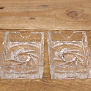May include: Two clear glass square-shaped ashtrays with a cut-glass design. The ashtrays have a small notch on one side.