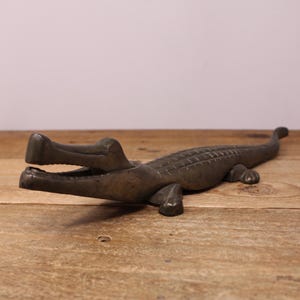 May include: A dark bronze-toned metal alligator figurine with an open mouth, textured scales along its back, and four short legs. The sculpture is elongated with a long tail extending toward the back, showcasing a weathered, metallic finish.