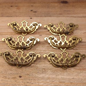 May include: A set of six brass drawer pulls featuring an ornate, openwork backplate with a curved, swinging handle. Each piece displays a metallic gold finish with intricate scrollwork details and a symmetrical design for furniture hardware.