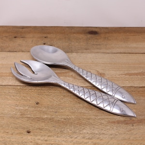 May include: A set of two silver-toned serving utensils shaped like fish. The utensils are a spoon and a fork, both with a textured surface.