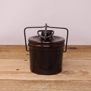 Peut inclure: Un pot en céramique cylindrique brun foncé avec un couvercle assorti et un mécanisme de poignée en fil de fer gris qui maintient le couvercle en place. Le récipient présente une finition lisse et brillante ainsi qu'un design fonctionnel.