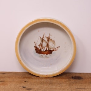 May include: A round ceramic dish with a white interior and a light orange rim. The center of the dish features a detailed illustration of a brown sailing ship with white sails on blue water. The surface has subtle speckles throughout the design.