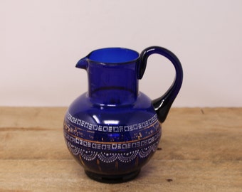 May include: A cobalt blue glass pitcher with a gold and white geometric design. The pitcher has a rounded body and a small spout.