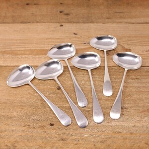 May include: Seven silver serving spoons with oval bowls and long, slender handles, arranged on a wooden surface. The spoons reflect light, and each handle features a small, engraved detail.