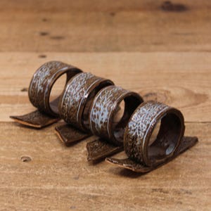 May include: Four ceramic napkin rings with a textured, brown exterior and a white and blue pattern. Each ring sits on a matching rectangular base. The rings are arranged in a curved line on a wooden surface.