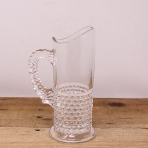 May include: Clear glass pitcher with a textured, hobnail-style base and a twisted handle. The pitcher has a curved spout and sits on a round base. The background is a light color, and the pitcher is on a wooden surface.