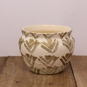 Peut inclure: Un pot en céramique blanche avec un motif géométrique doré. Le pot a une forme arrondie et une surface texturée.