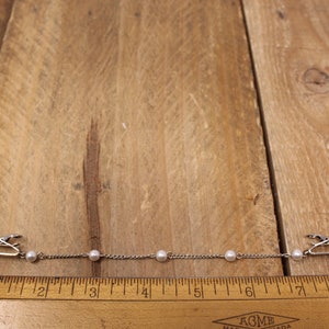 May include: A silver chain with five white pearls and two silver clips on either end. The chain is about 18 cm long and is measured against a wooden ruler.