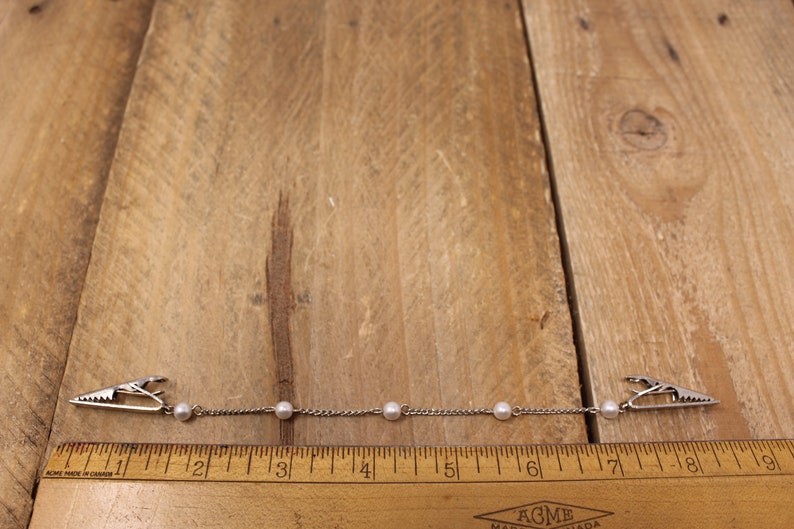 May include: A silver chain with five white pearls and two silver clips on either end. The chain is about 18 cm long and is measured against a wooden ruler.