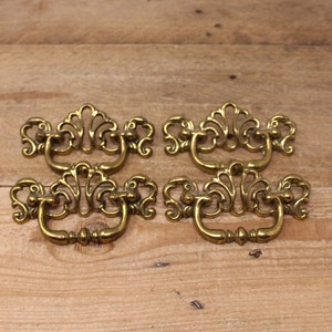 May include: Four ornate brass drawer pulls with a decorative, floral design. The pulls have a loop handle and are in a vintage style.