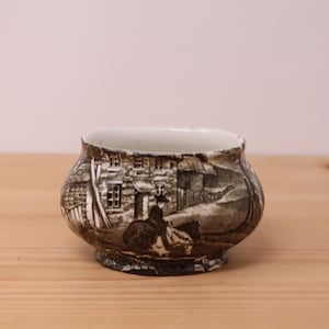 May include: A small, round ceramic bowl featuring a dark brown and white transferware pattern. The design depicts a rural scene with a stone cottage, a fence, and a person riding a horse. The interior of the bowl is white, and it has a flared rim.