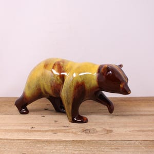 May include: A ceramic bear figurine in a walking pose. The bear has a glossy finish with a colour scheme of brown, yellow, and reddish-brown. The figurine is placed on a wooden surface.
