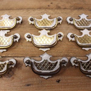 May include: Nine decorative cabinet pulls in white and gold. Each pull has a white frame, gold diamond-patterned insert, and bronze-coloured accents. The pulls are displayed on a wooden surface.