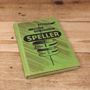 May include: A vintage green "SPELLER" book with black and white graphics. The book cover reads "The Pupils' Own Vocabulary" and "Book 1 Grades II-III". The book is on a wooden surface.