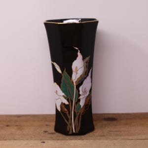 May include: A tall, hexagonal black vase featuring a painted design of white calla lilies with green leaves and gold-toned outlines. The rim of the vase is finished with a thin gold-coloured border, providing a distinct contrast against the dark surface.