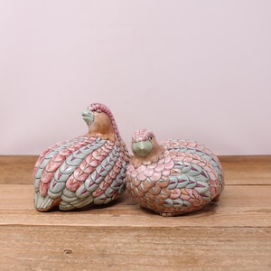 May include: Two ceramic bird figurines. The birds have a light brown head and a body covered in a pattern of pink, blue, and white feathers. The figurines are placed on a wooden surface.