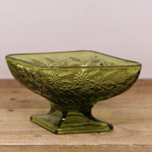 Peut inclure: Bol en verre vert sur pied, avec un dessus carré et un motif floral en relief. Le bol a une base carrée courte et une tige décorative. Le bol est probablement vintage et convient pour servir des collations ou exposer de petits objets.