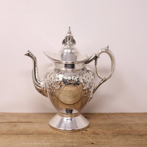 May include: A silver teapot with an ornate design and a lid with a finial. The teapot has a round body with a flared spout and a handle.