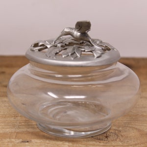 May include: A clear glass jar with a silver lid featuring a hummingbird design. The lid has a decorative openwork pattern.