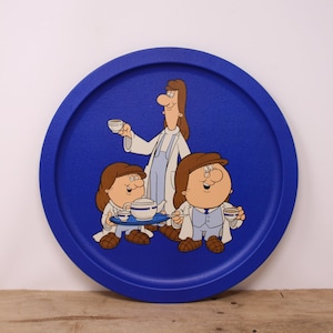 May include: A round, cobalt blue serving tray with a cartoon illustration. The illustration depicts a woman and two children, all dressed in white lab coats, serving tea. The tray has a textured surface and a raised edge.