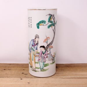 May include: A cylindrical white ceramic vase with a hand-painted scene of two figures under a tree. The vase features a gold rim and a painted landscape with figures in traditional clothing. The scene includes a tree with green and orange foliage.