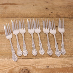 May include: Eight silver cake forks with ornate handles. The forks are arranged in a row on a wooden surface.
