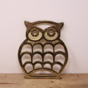 May include: Gold metal owl-shaped hot pad with a cutout design. The owl has large, round eyes and a detailed feather pattern.
