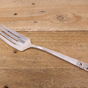 May include: A silver fork with an ornate handle. The fork has a polished finish and is lying on a wooden surface.