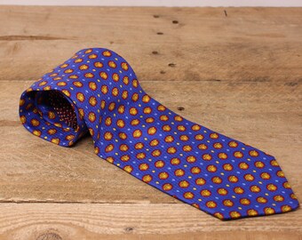 May include: A blue tie with a repeating pattern of yellow and red circles and blue diamonds. The tie is made of a textured fabric and is displayed on a wooden surface. The tie is a classic accessory.