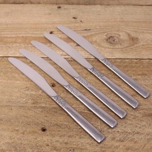 May include: Five silver dinner knives with a decorative pattern on the handles. The knives are arranged in a row on a wooden surface.