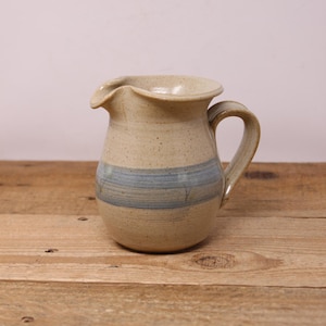 May include: A small, beige ceramic jug with a blue and white stripe design. The jug has a wide, rounded base and a flared lip.