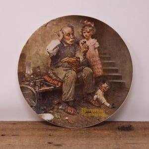 May include: Circular decorative plate with a vintage illustration of a cobbler, a young girl, and a doll. The cobbler is seated, examining a shoe. The girl holds a flower. The plate has a brown and beige colour scheme, with the artist's signature.