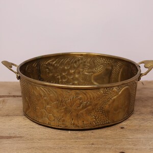 May include: A round, brass planter with two handles. The planter has an embossed design of flowers and leaves.