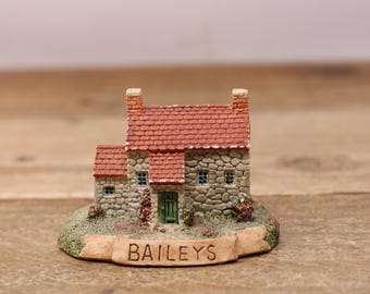 May include: A miniature stone cottage with a red tiled roof and a green door. The cottage is surrounded by greenery and has a sign that reads "BAILEYS".