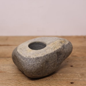 May include: A grey stone candle holder with a round opening in the top. The holder is sitting on a wooden surface.