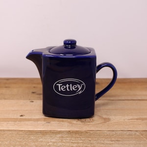 Peut inclure: Théière en céramique bleu marine de forme carrée avec une anse incurvée. Elle est dotée d'un couvercle et d'un bec verseur. Le mot "Tetley" est imprimé en blanc sur le devant. Posée sur une surface en bois.
