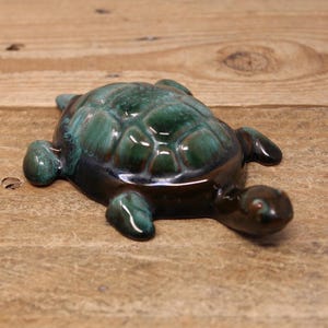 May include: A ceramic turtle figurine with a green and brown glaze. The turtle is lying on its belly with its head turned to the side.