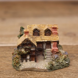 May include: A miniature cottage figurine with a thatched roof, brown wooden walls, and red brick accents. The cottage has two windows with white panes and a small door. The figurine is surrounded by green foliage and small purple flowers.