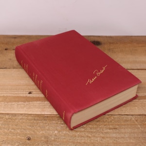 May include: A hardcover book with a red cover and gold lettering. The book has a signature in gold on the cover. The pages are cream-colored, and the book is resting on a wooden surface. The book's title is partially visible.