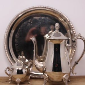 Peut inclure: Un plateau de service en argent avec une théière, un pot à crème et un sucrier en argent. Le plateau a une bordure décorative et la théière a un couvercle et un bec.