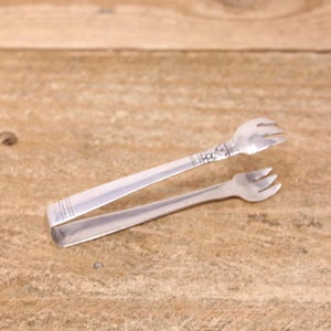 May include: Silver sugar tongs with a floral handle design. The tongs have fork-shaped ends for picking up sugar cubes. The tongs are open and resting on a wooden surface.