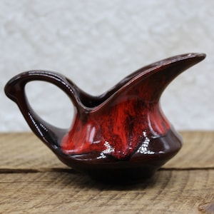 May include: A small ceramic creamer pitcher featuring a glossy finish with a deep dark brown base and a vibrant red glaze accent on the upper body. The pitcher has a curved handle and a pointed pouring spout, showcasing a smooth, reflective surface.