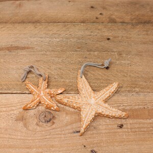 May include: Two starfish ornaments in off-white and orange with a textured finish. Each has a grey string loop for hanging. They are displayed on a rustic wooden surface.