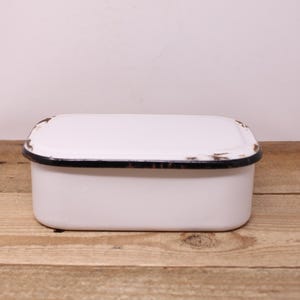 May include: A white rectangular enamelware container with a lid. The container has a black rim with a distressed, aged appearance. The piece is set on a wooden surface, suggesting a vintage or rustic aesthetic.